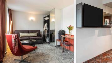 A modern hotel room with a sofa, a red chair, a small table, a desk, a mirror, and a TV.