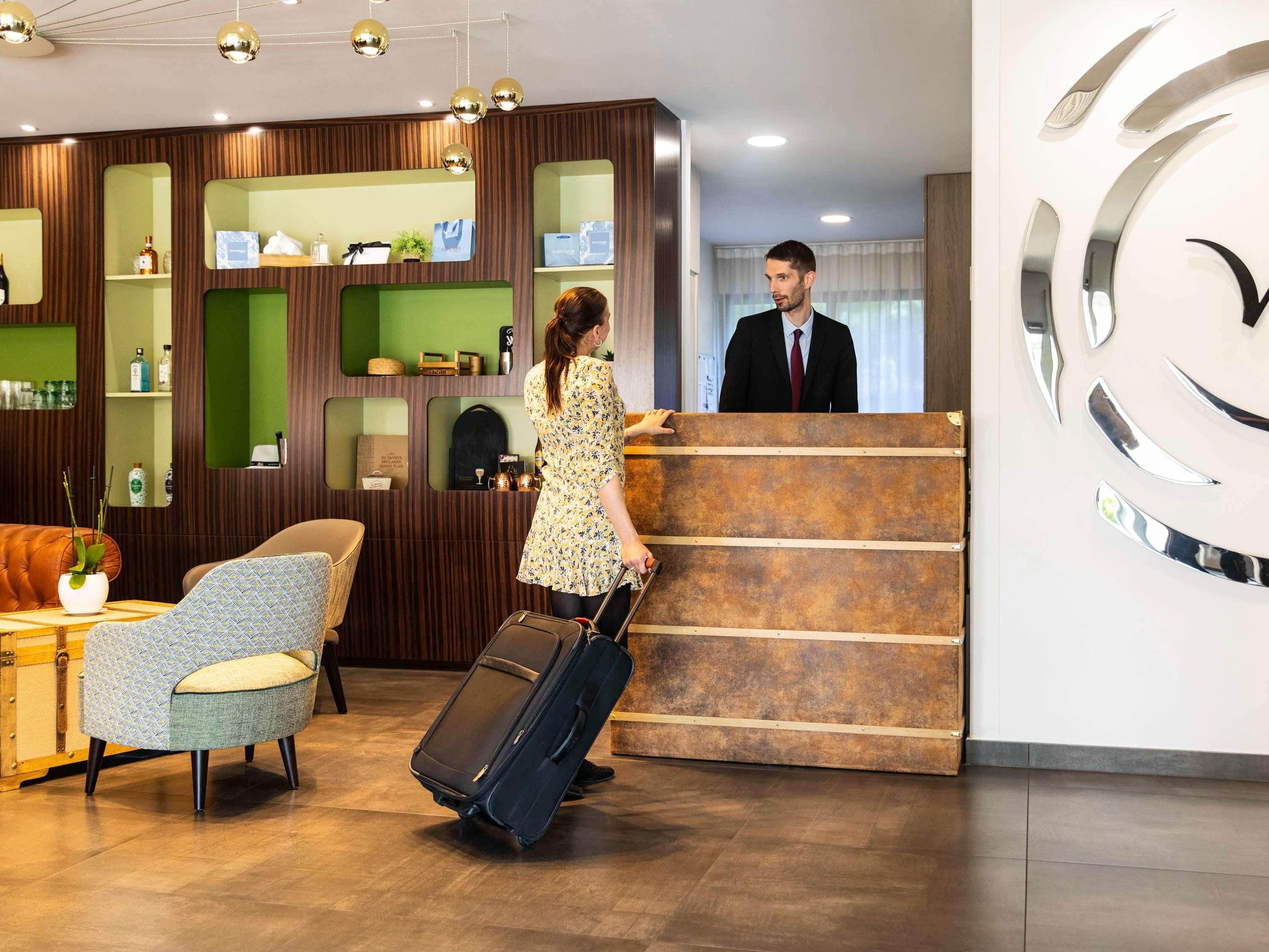Un espace d’accueil d’hôtel comprenant un comptoir de réception en bois, un mur végétal agrémenté d’étagères, un canapé en cuir et un fauteuil à motifs.