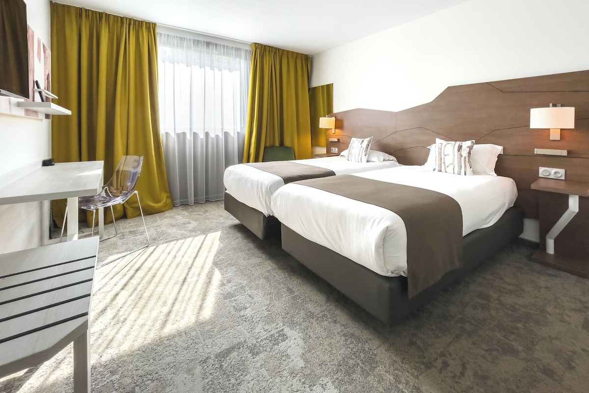 A hotel room with two beds, a desk, a chair, and a large window with curtains.