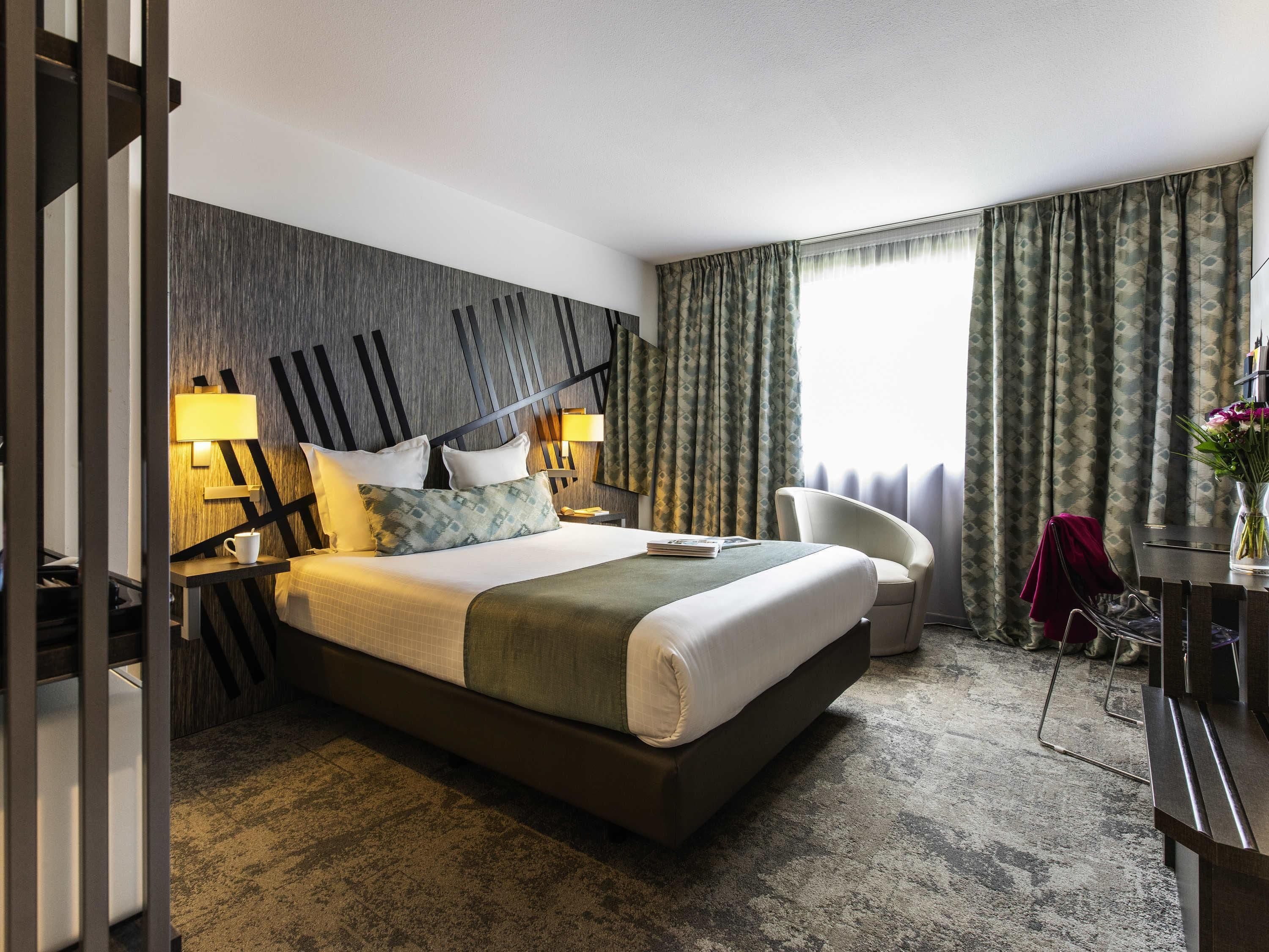 Een hotelkamer met een groot bed, twee bedlampen, een stoel, een bureau en een raam met gordijnen.