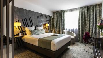 Een hotelkamer met een groot bed, twee bedlampen, een stoel, een bureau en een raam met gordijnen.