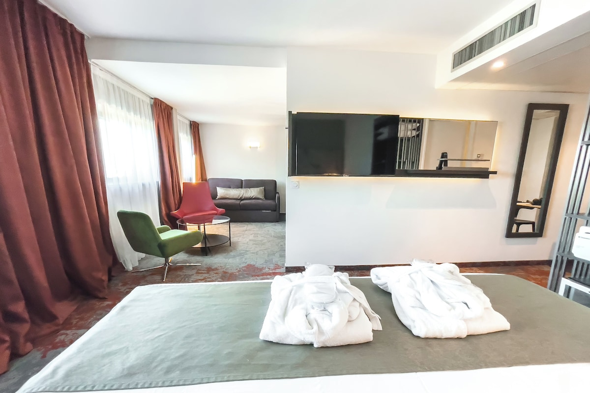 A hotel room with a bed, a green armchair, a red chair, a sofa, a TV, and a mirror.