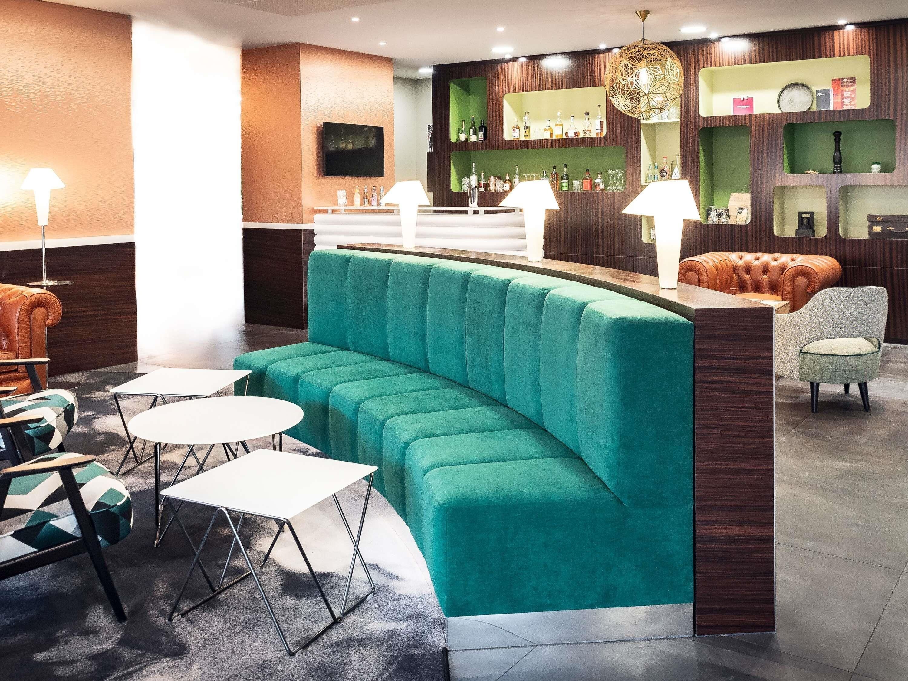 A modern bar area with a teal seating section, white tables, and a variety of bottles on shelves.
