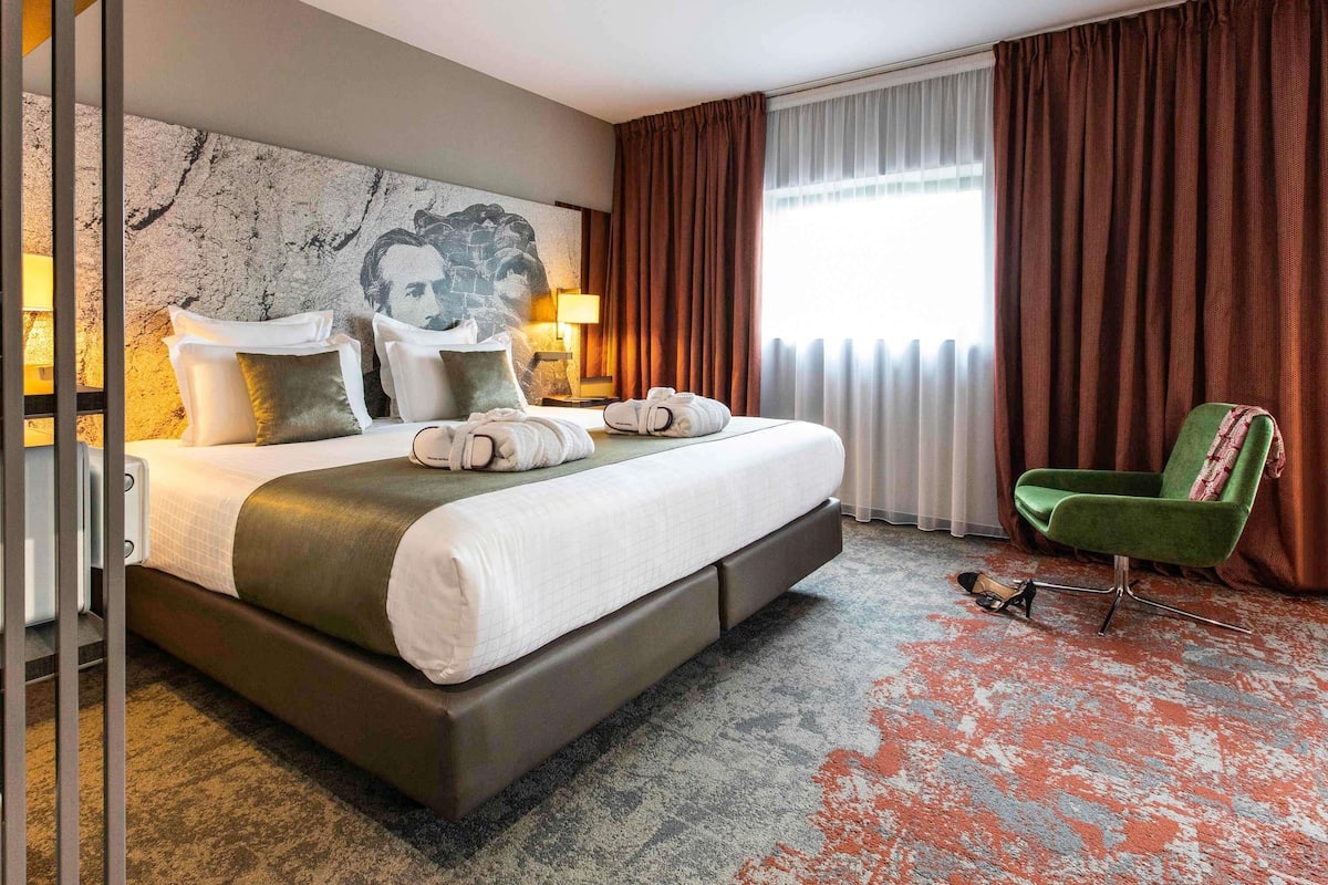 Ein Hotelzimmer mit einem großen Bett, einem grünen Sessel, einem Marmorkopfschild und Blick ins Freie.