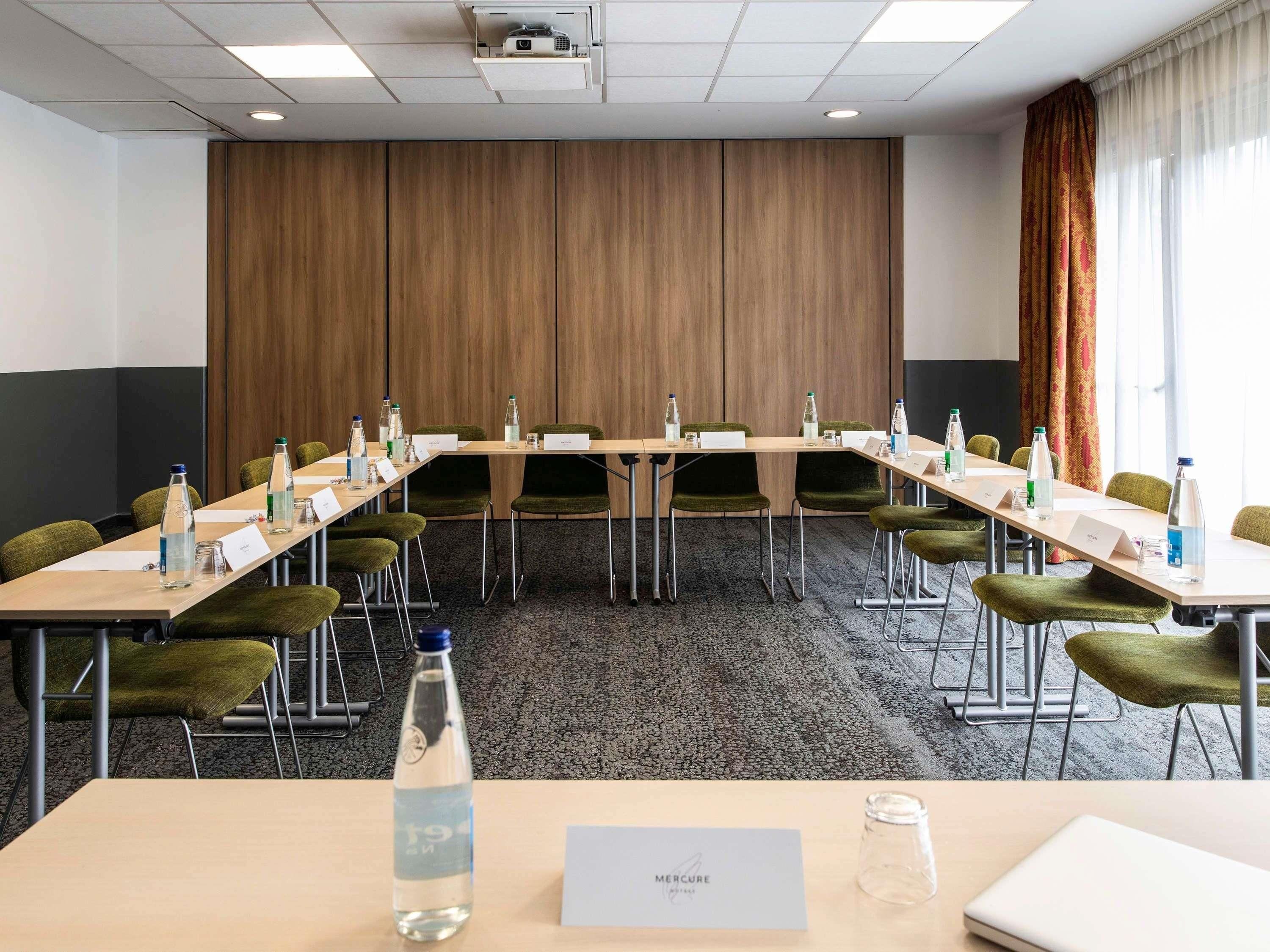 A conference room with a long table, green chairs, nameplates, water bottles, and a projector on the ceiling.