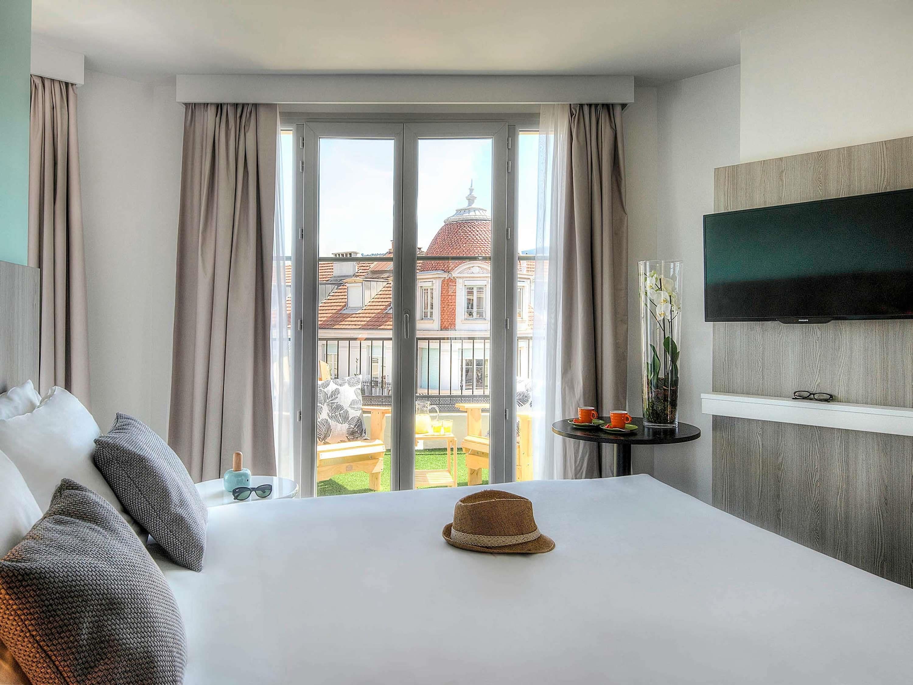 A hotel room with a bed, a TV mounted on the wall, a small table with a vase and two cups, and a view of a building through a window.