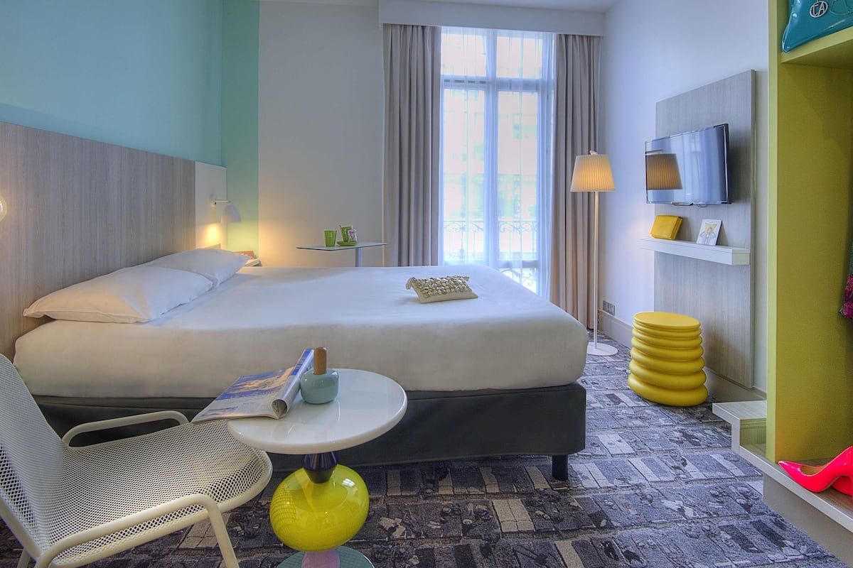 A hotel room with a bed, a chair, a small table, a lamp, a TV, and a shelf with a handbag.