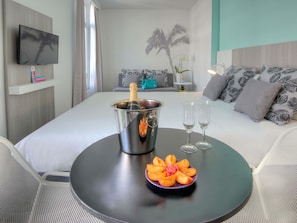 A hotel room with a bed, a small round table with a champagne bucket, two glasses, and a fruit arrangement.