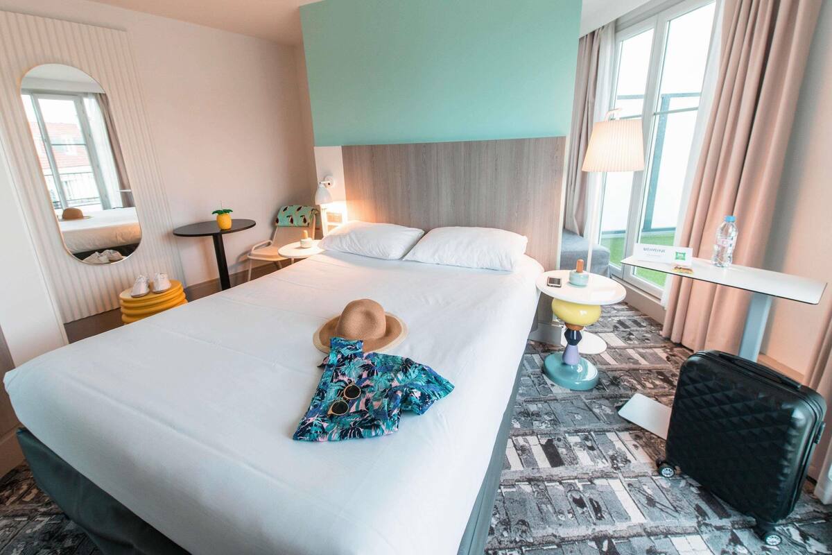 A hotel room with a bed, a bedside table, a lamp, a chair, a mirror, and a suitcase.