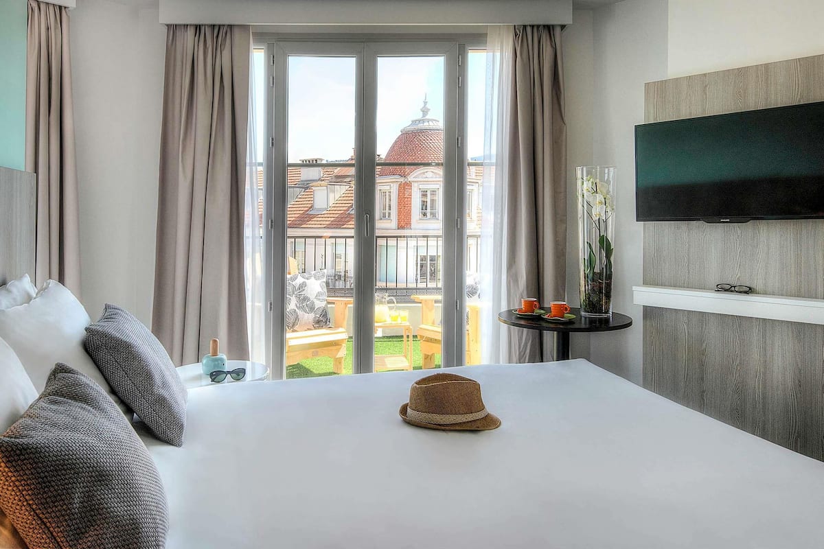 Ein Hotelzimmer mit Bett, einem an der Wand befestigten Fernseher, einem kleinen Tisch mit Vase und zwei Tassen sowie Blick auf ein Gebäude durch ein Fenster.