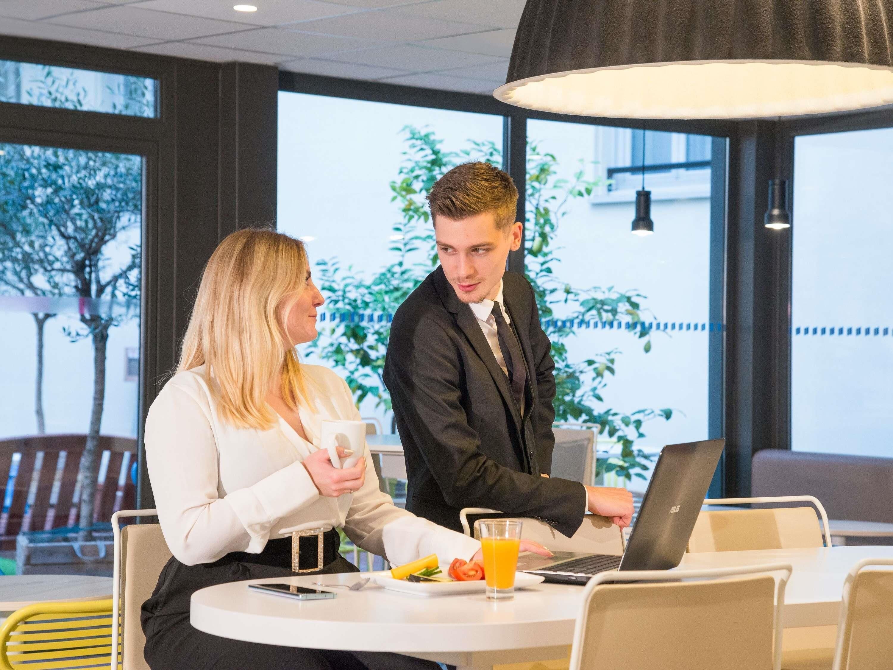 Deux personnes sont assises autour d’une table ronde. Sur la table, on peut voir un ordinateur portable, un verre de jus d’orange et une assiette de nourriture.