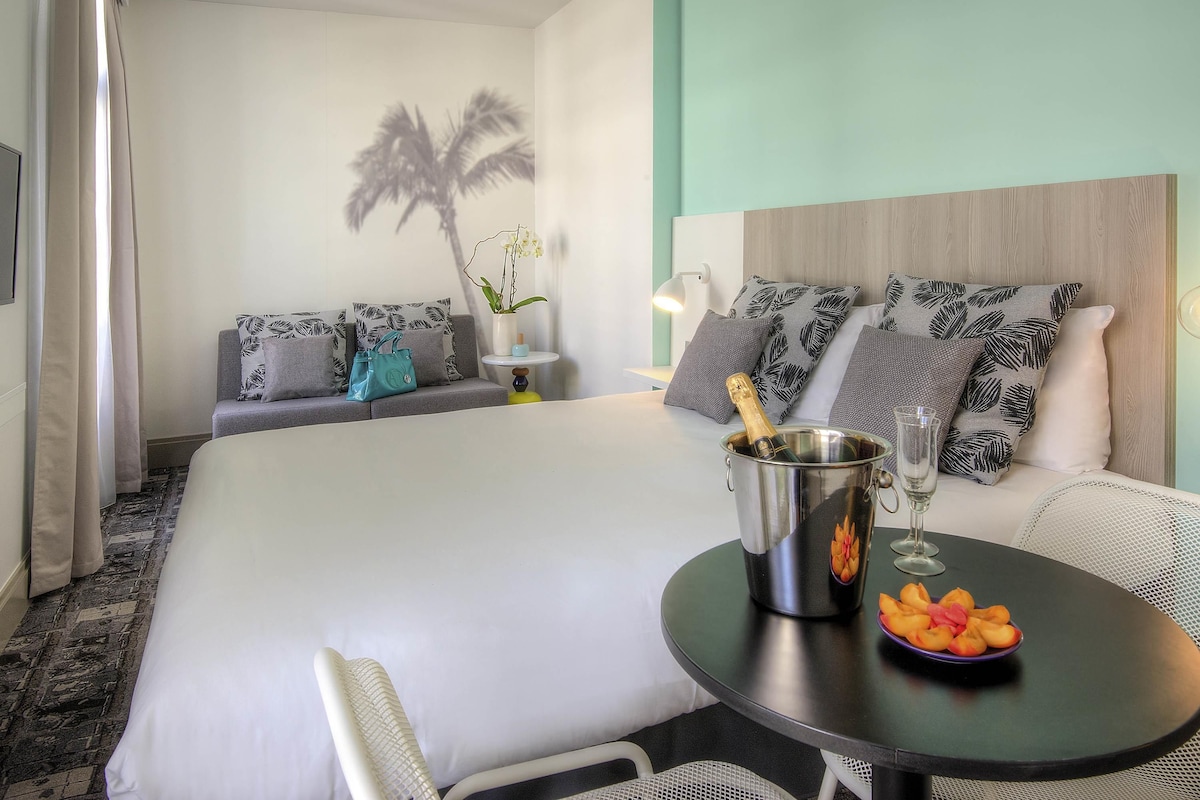 A hotel room with a bed, a small table with fruit and a bottle, a sofa with patterned cushions, and a wall with a palm tree shadow.