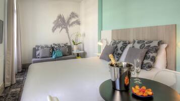 Une chambre d’hôtel avec un lit, une petite table sur laquelle se trouvent des fruits et une bouteille, un canapé avec des coussins à motifs et un mur sur lequel se projette l’ombre d’un palmier.