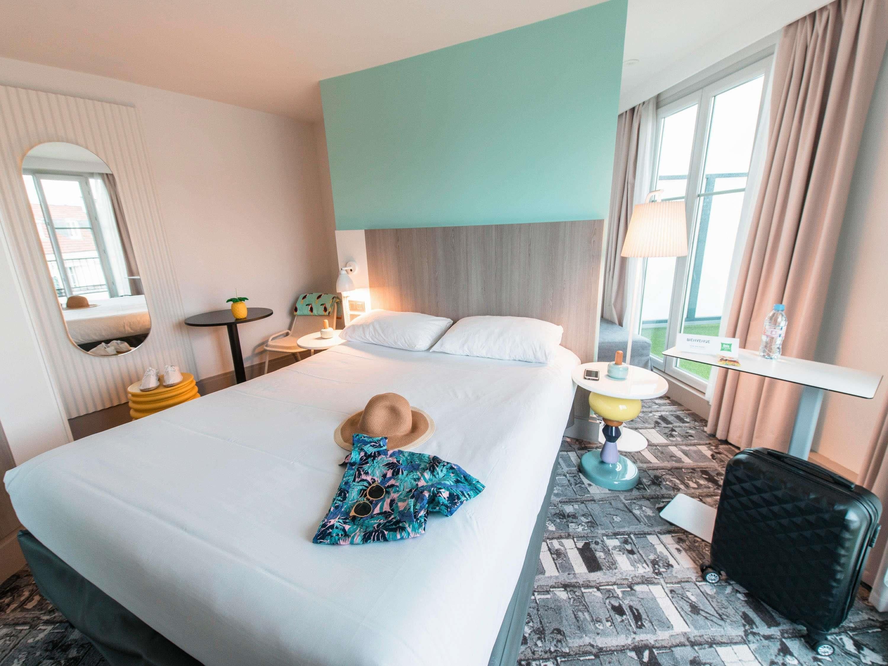 Une chambre d’hôtel avec un lit, une table de chevet, une lampe, une chaise, un miroir et une valise.