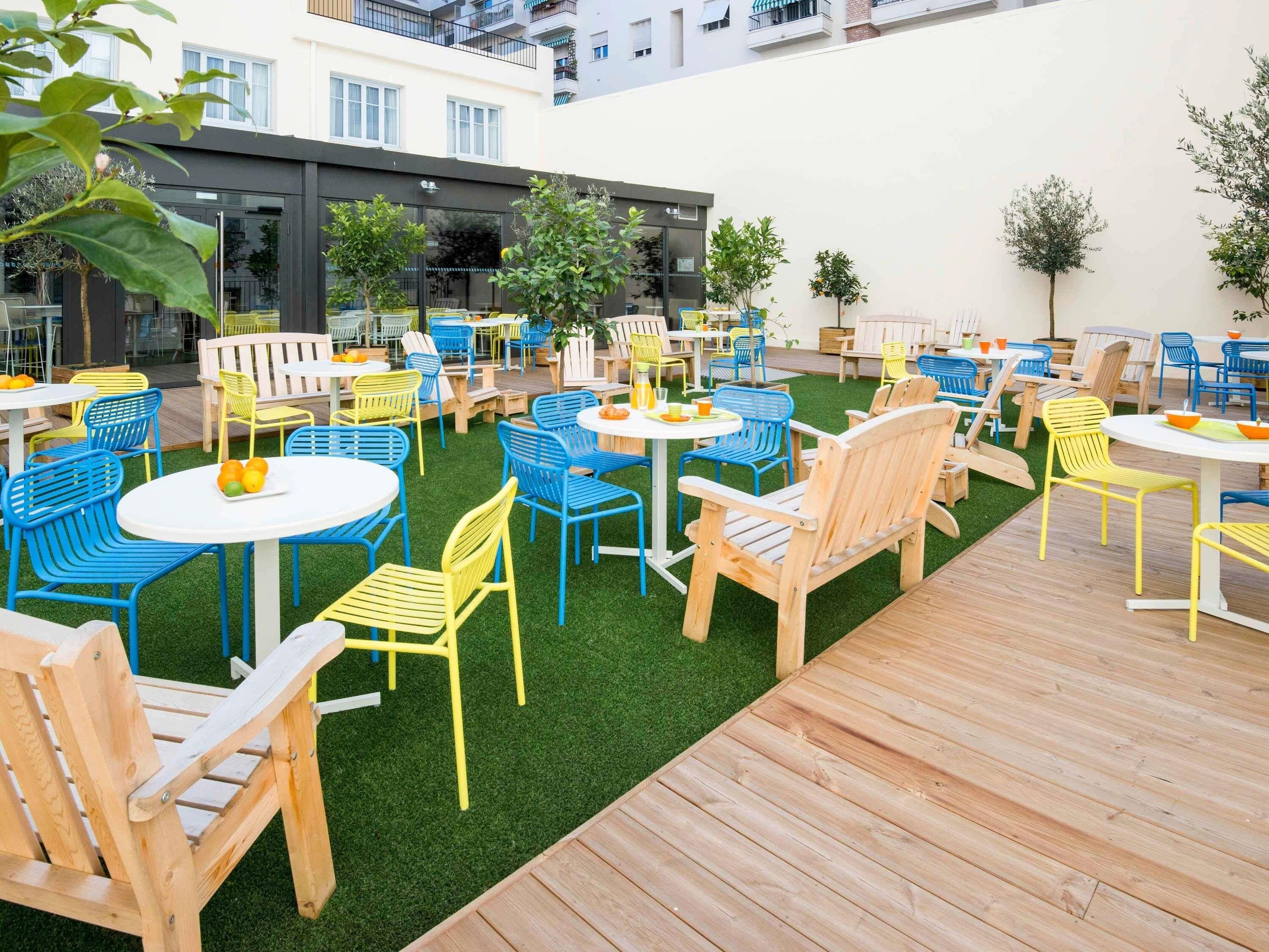 An outdoor seating area with multiple tables and chairs, surrounded by greenery and a wooden deck.