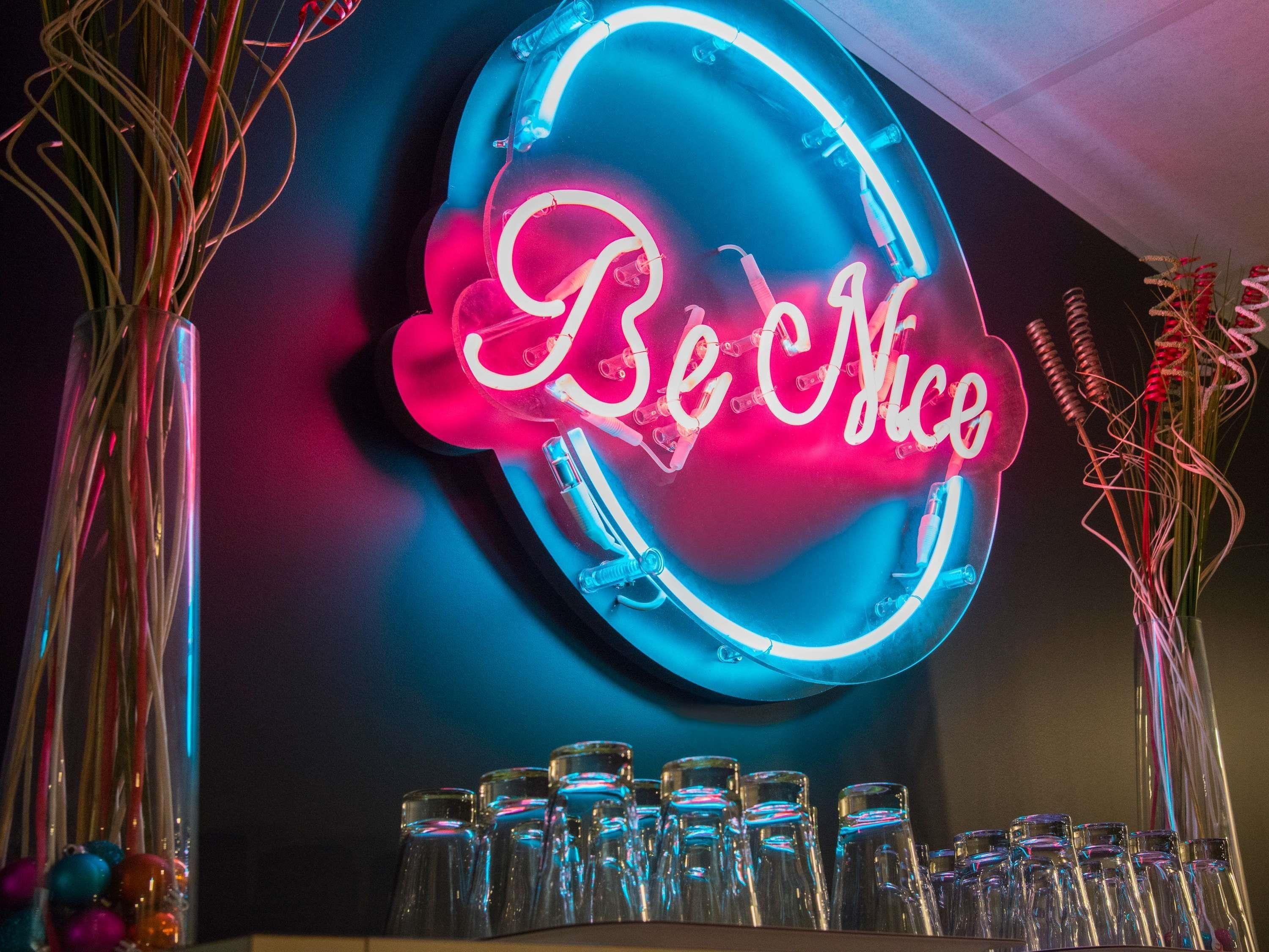 Un letrero de neón con el texto 'Be Nice' en un diseño circular, rodeado de botellas de vidrio transparentes.