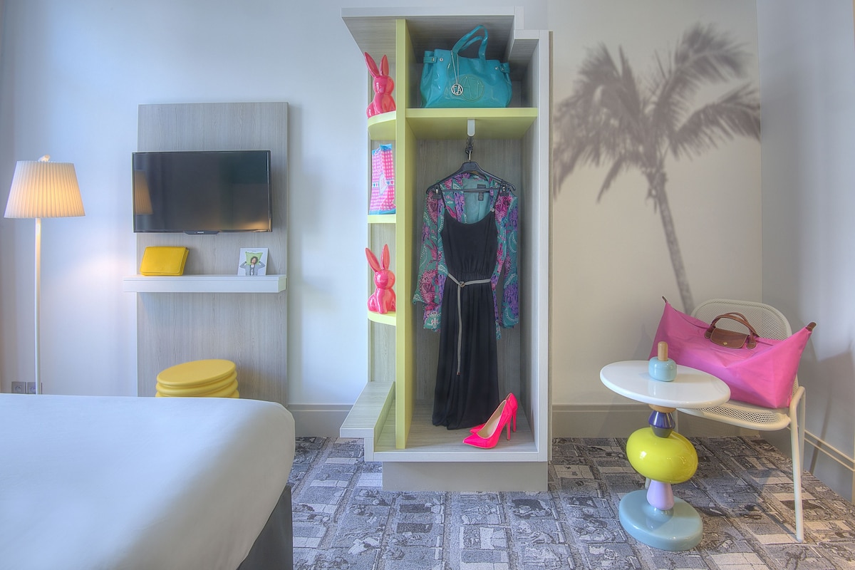 A hotel room with a bed, a TV, a wardrobe with hanging clothes, a small table with a lamp, and a palm tree decoration.