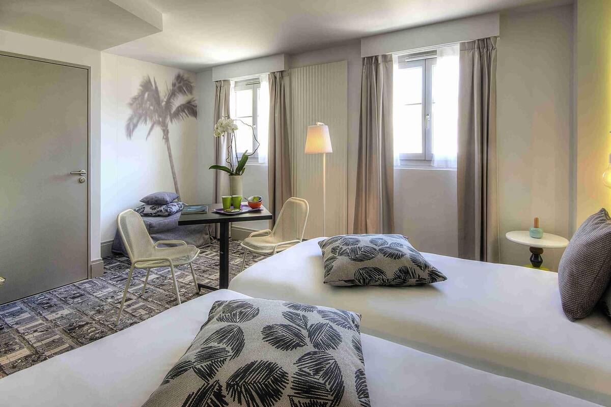 Ein Hotelzimmer mit zwei Betten, einer Sitzecke mit einem Sessel, einem kleinen Tisch mit einer Lampe und Blick ins Freie durch das Fenster.