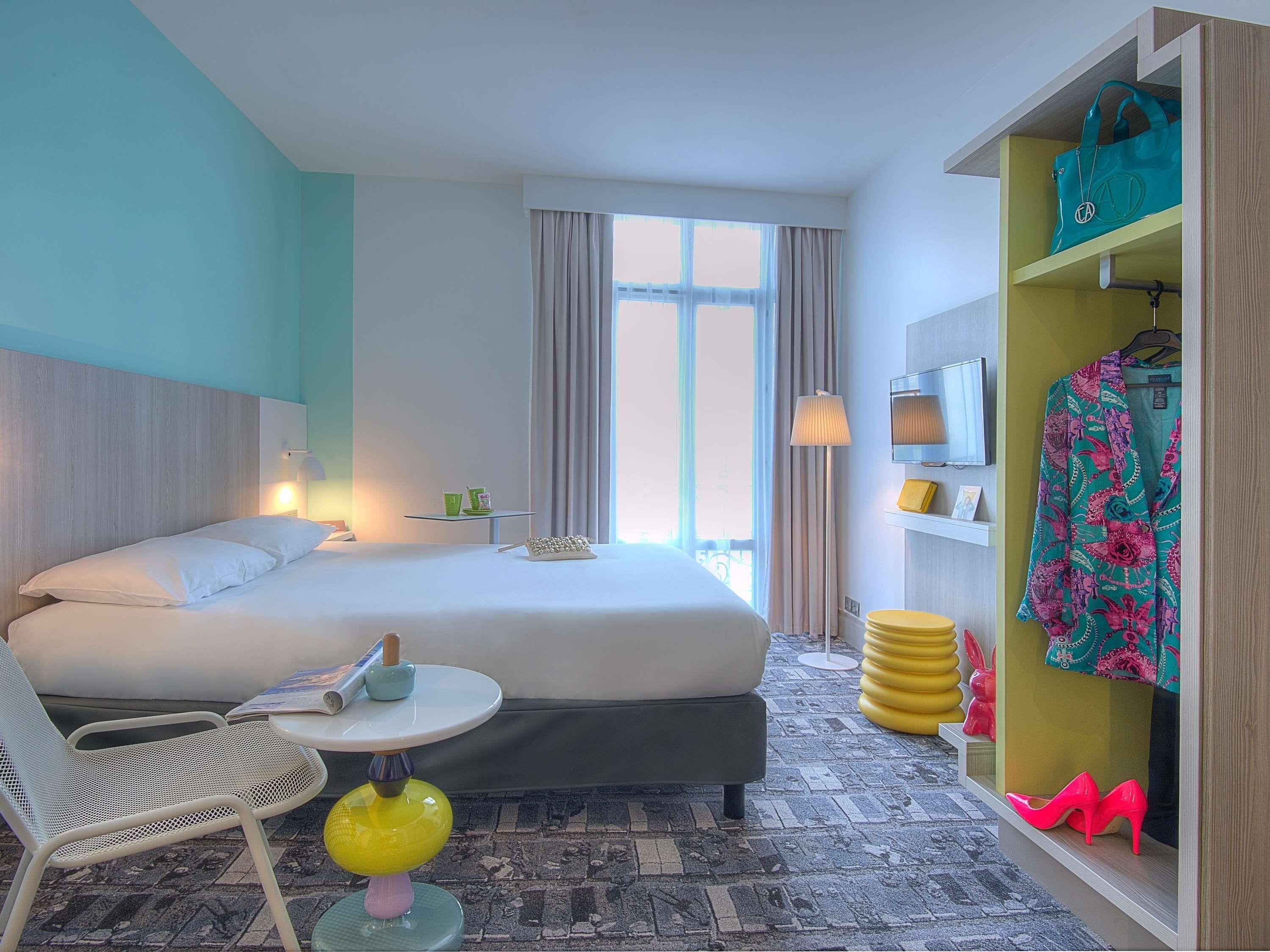 A hotel room with a bed, a chair, a small table, a lamp, a wardrobe, and a window with curtains.