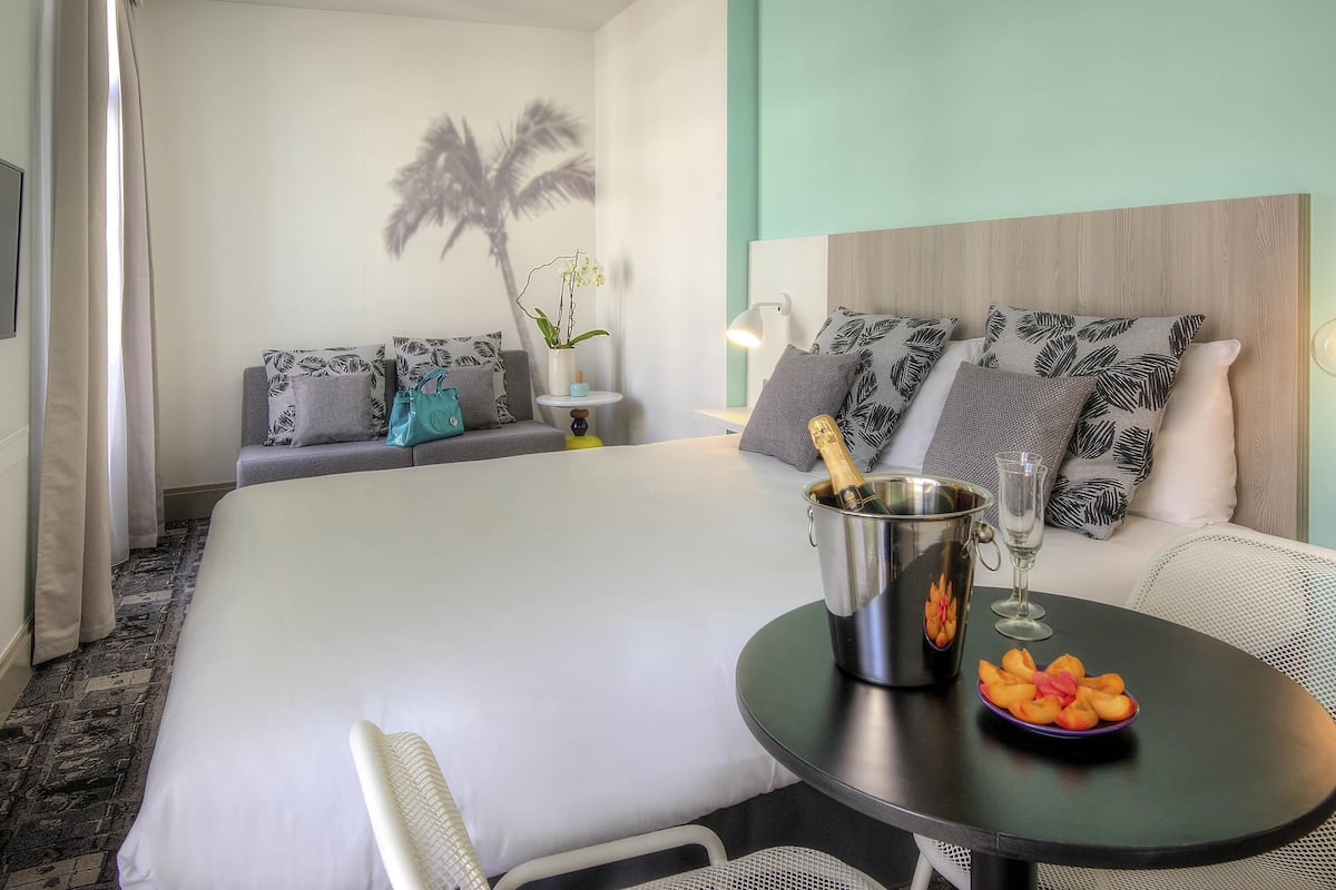 Ein Hotelzimmer mit Bett, einem kleinen Tisch mit Obst und einer Flasche, einer Couch mit gemusterten Kissen und einer Wand mit Palmen-Silhouette.