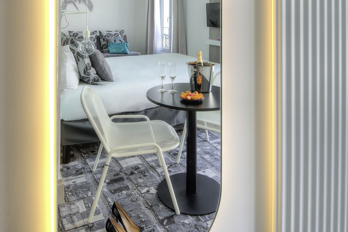 A modern hotel room with a bed, a small round table with a bottle and glasses, a chair, and a pair of shoes on the floor.