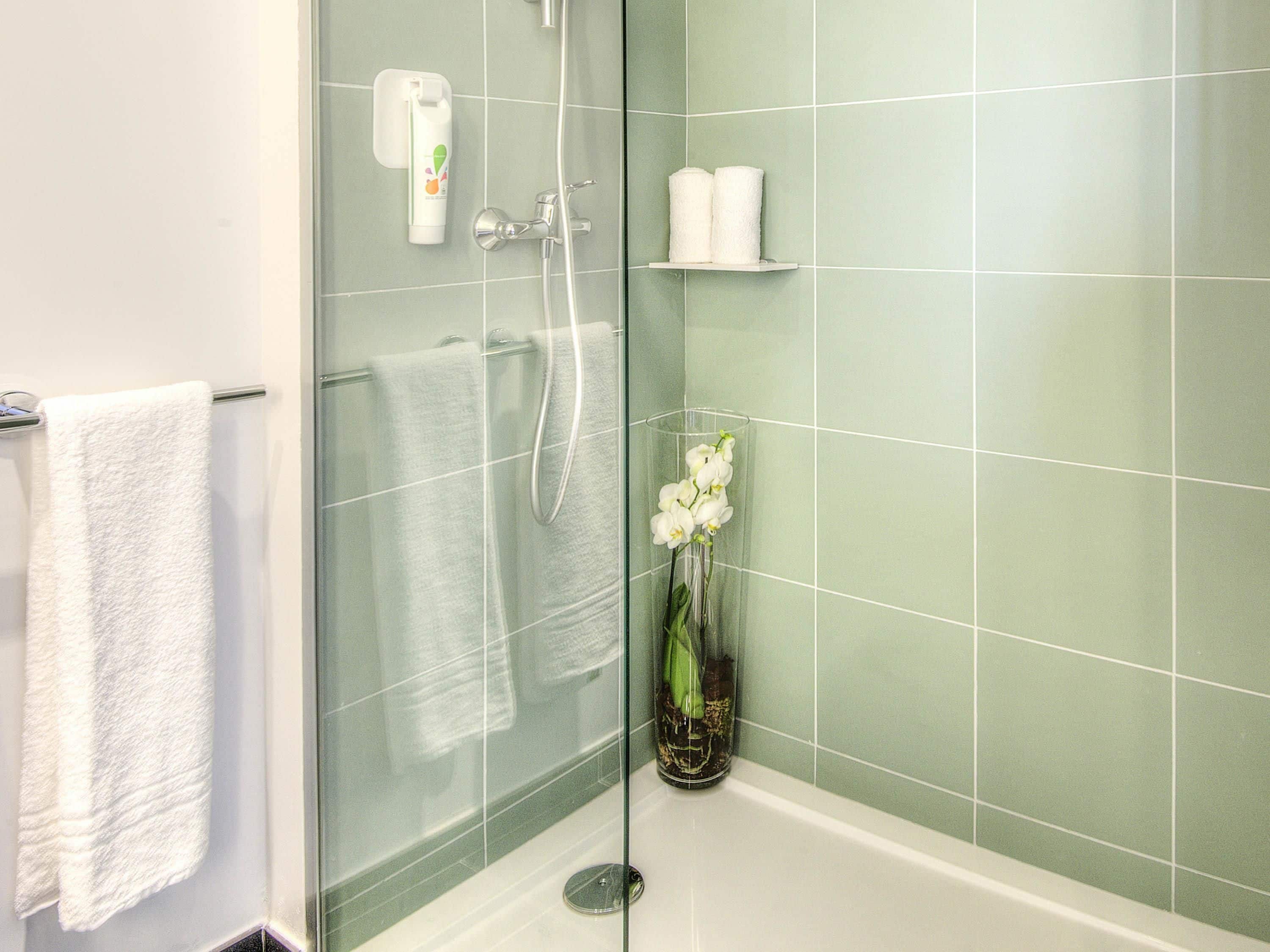 Un baño con cabina de ducha de vidrio, una toalla blanca colgada en un perchero, un florero de vidrio con flores blancas y un estante con dos toallas blancas.