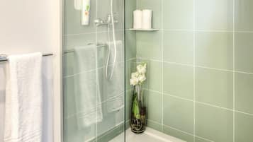 Un baño con cabina de ducha de vidrio, una toalla blanca colgada en un perchero, un florero de vidrio con flores blancas y un estante con dos toallas blancas.