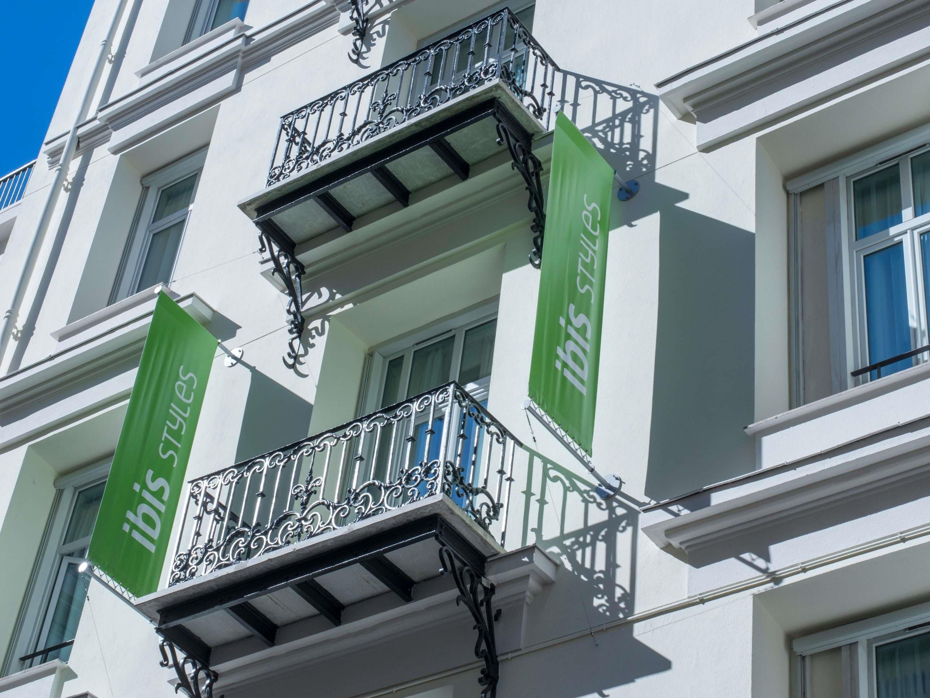 Un edificio con balcón que presenta barandillas de metal negro ornamentadas y un estandarte verde con el texto 'ibis styles'.