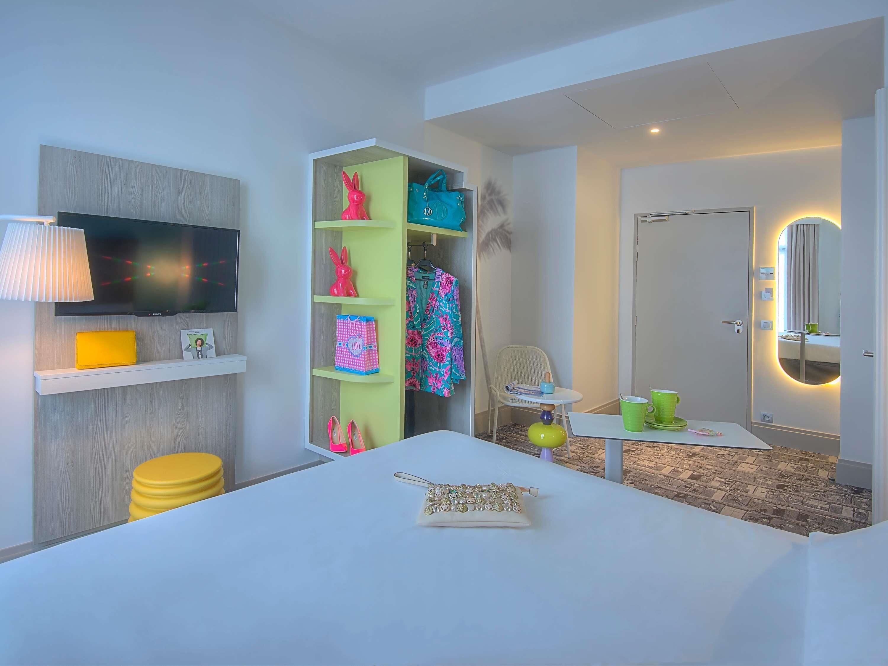 A modern hotel room with a flat-screen TV, a small table with a lamp, a vanity with a mirror, and a seating area with a chair and table.
