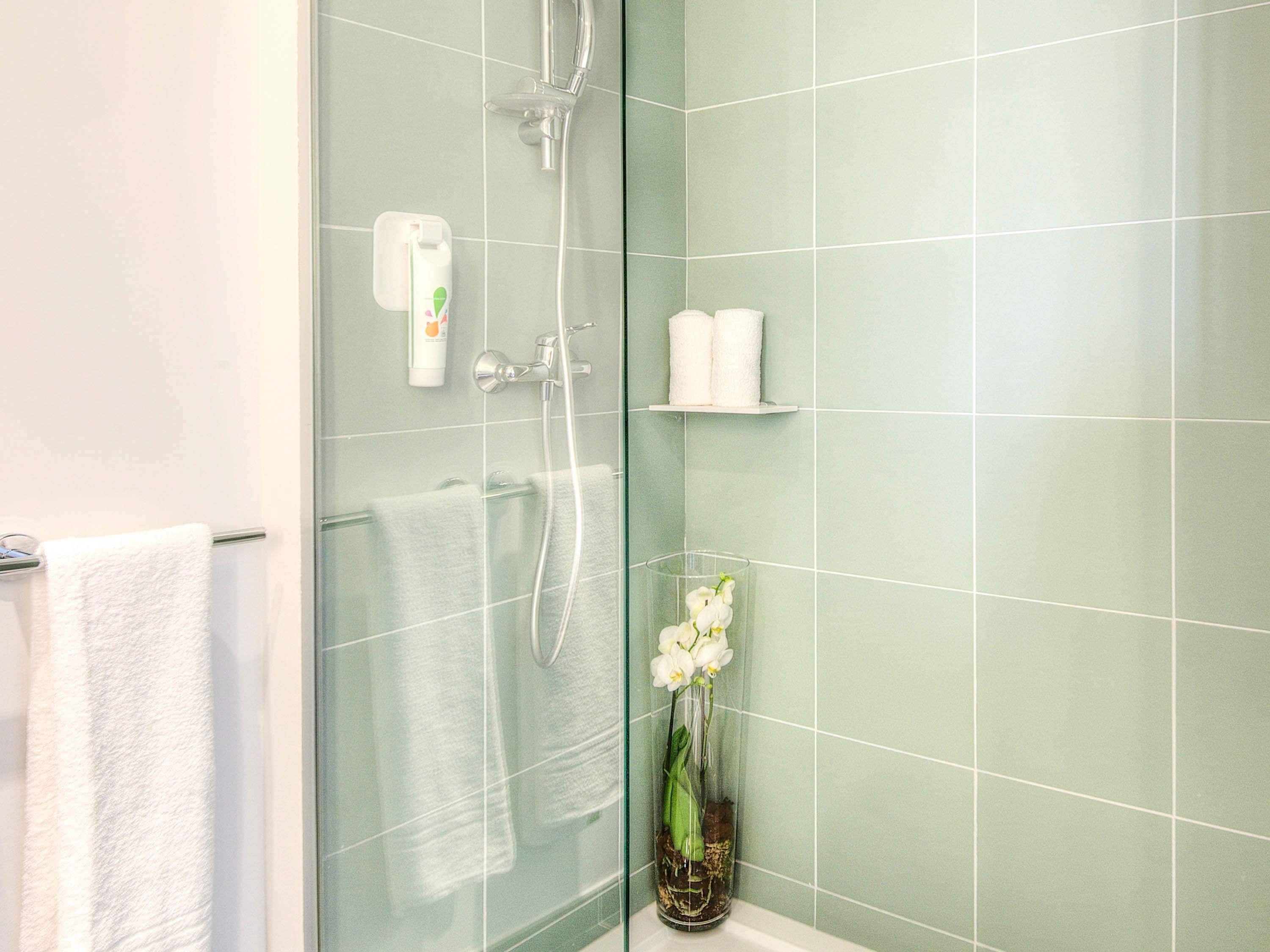 Un baño con cabina de ducha de vidrio, toallas blancas, un florero de vidrio con flores y un estante con artículos de aseo.