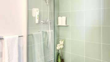 Un baño con cabina de ducha de vidrio, toallas blancas, un florero de vidrio con flores y un estante con artículos de aseo.