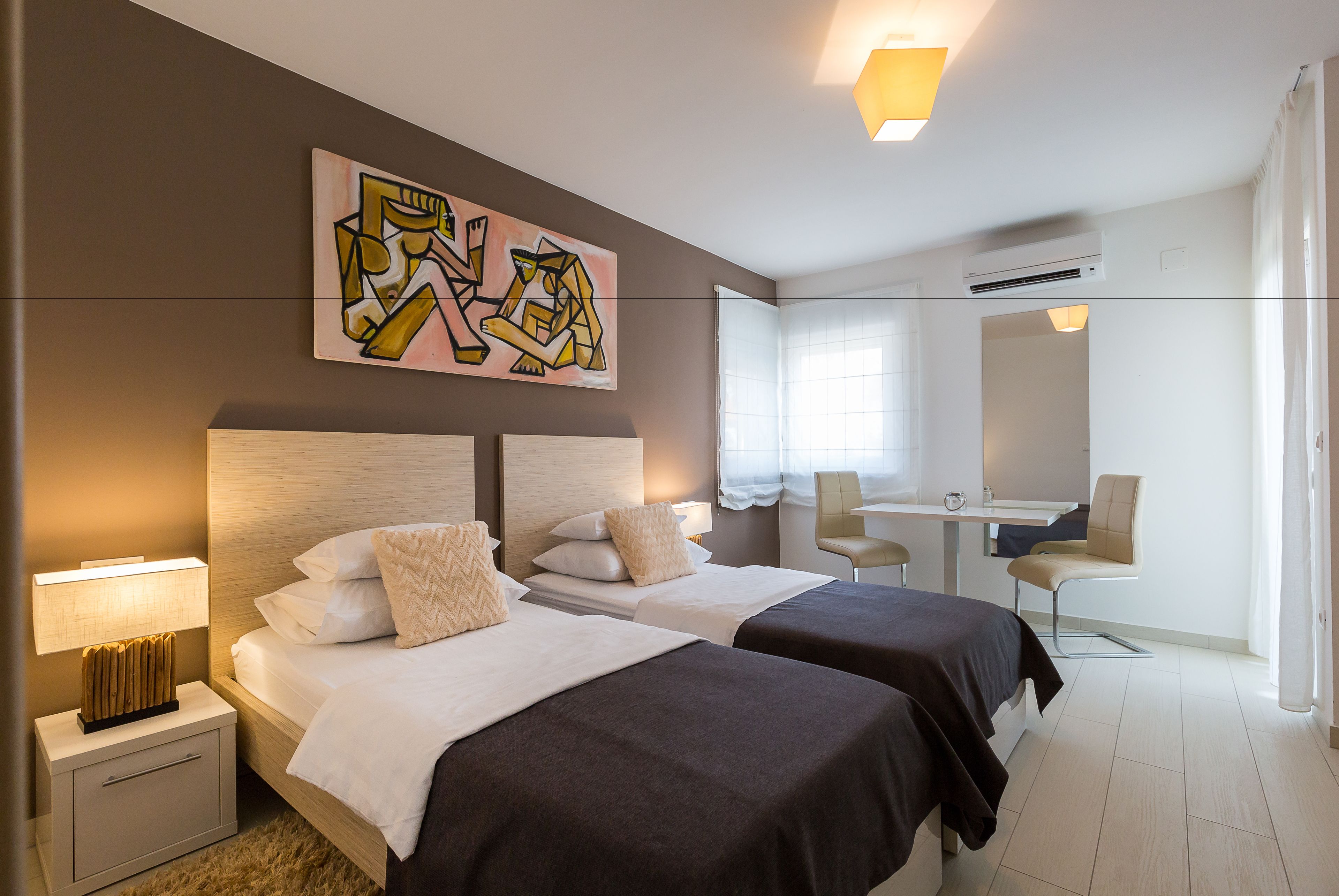 A hotel room with two beds, a desk, and a chair. There is a painting on the wall and an air conditioning unit above the desk.