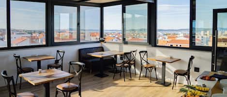 A restaurant with a high view of a cityscape, wooden tables and chairs, and a fruit bowl on the table.
