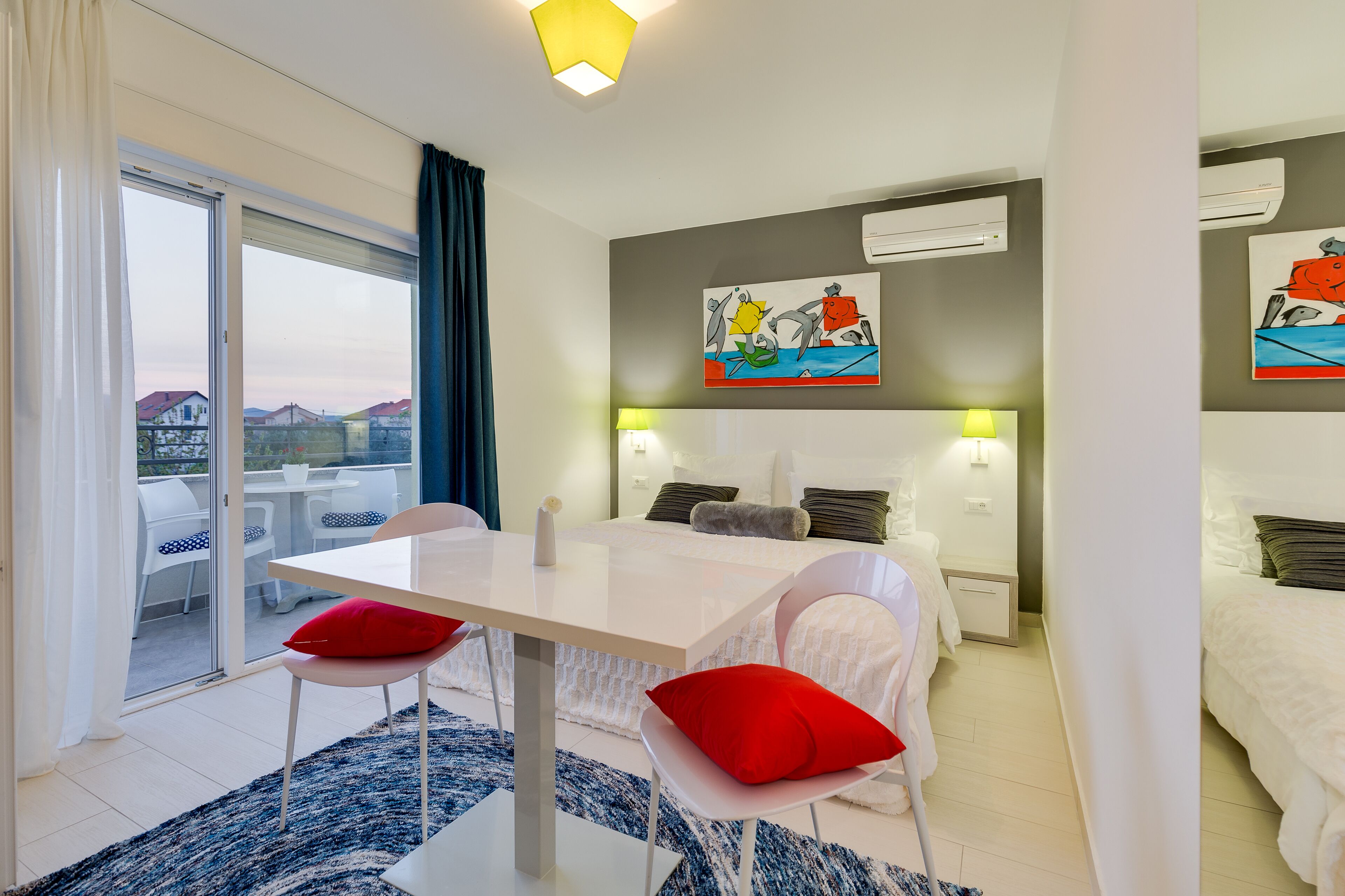 Una habitación de hotel moderna con comedor, una cama con ropa de cama blanca, almohada roja y un cuadro de delfines en la pared.