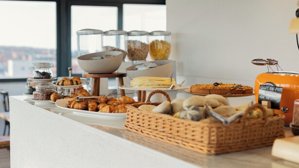 En buffet med forskellige bagværk, blandt andet croissanter, kager og brød, anrettet på en disk med byudsigt i baggrunden.