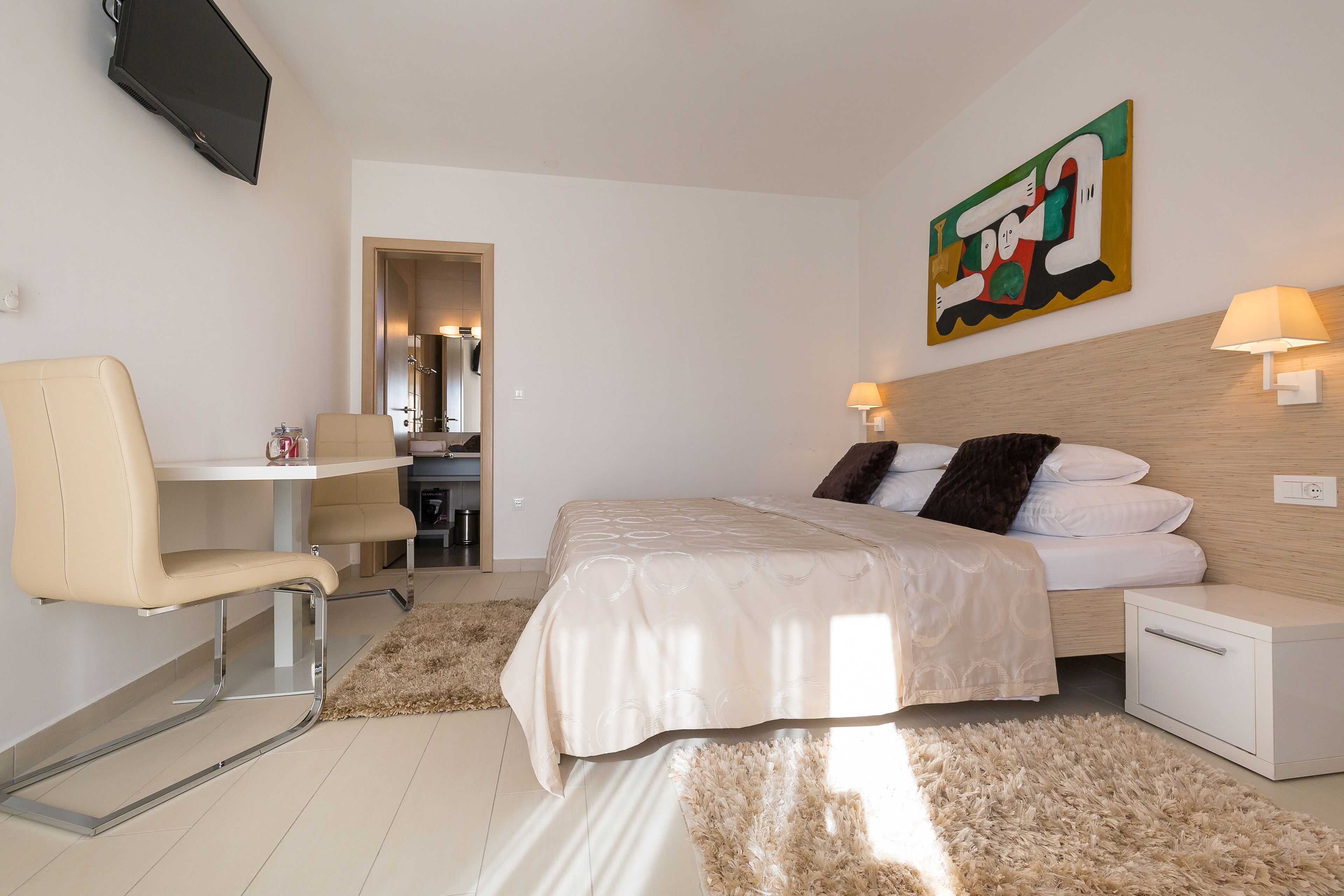 A modern hotel room with a bed, a desk, a chair, a TV, and a painting on the wall.