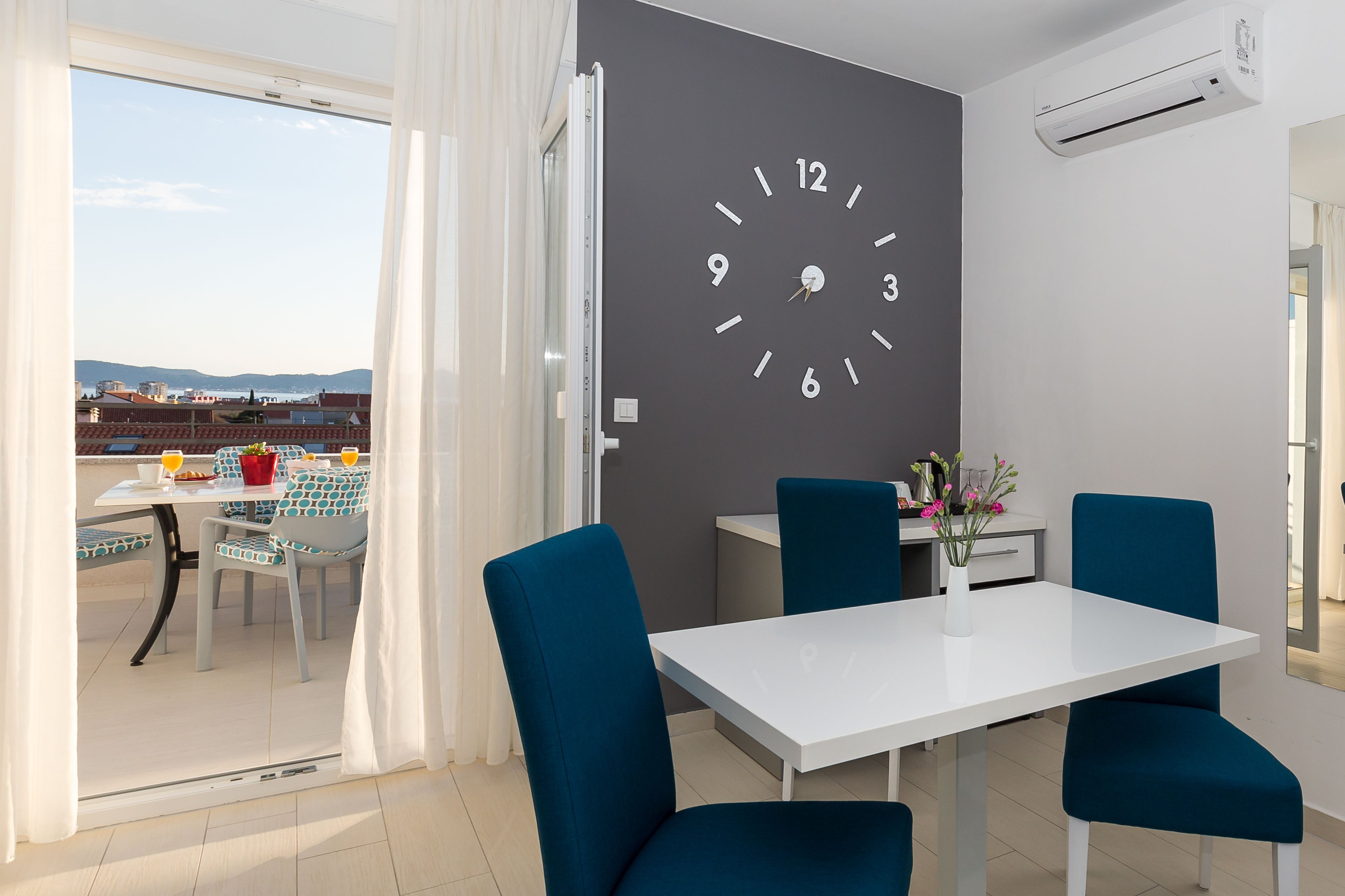 Ein moderner Essbereich mit einem weißen Tisch, blauen Stühlen, einer großen Wanduhr und Blick durch eine Glastür.