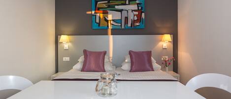 A modern hotel room with a bed, two bedside lamps, a small table with a vase, and a painting on the wall.