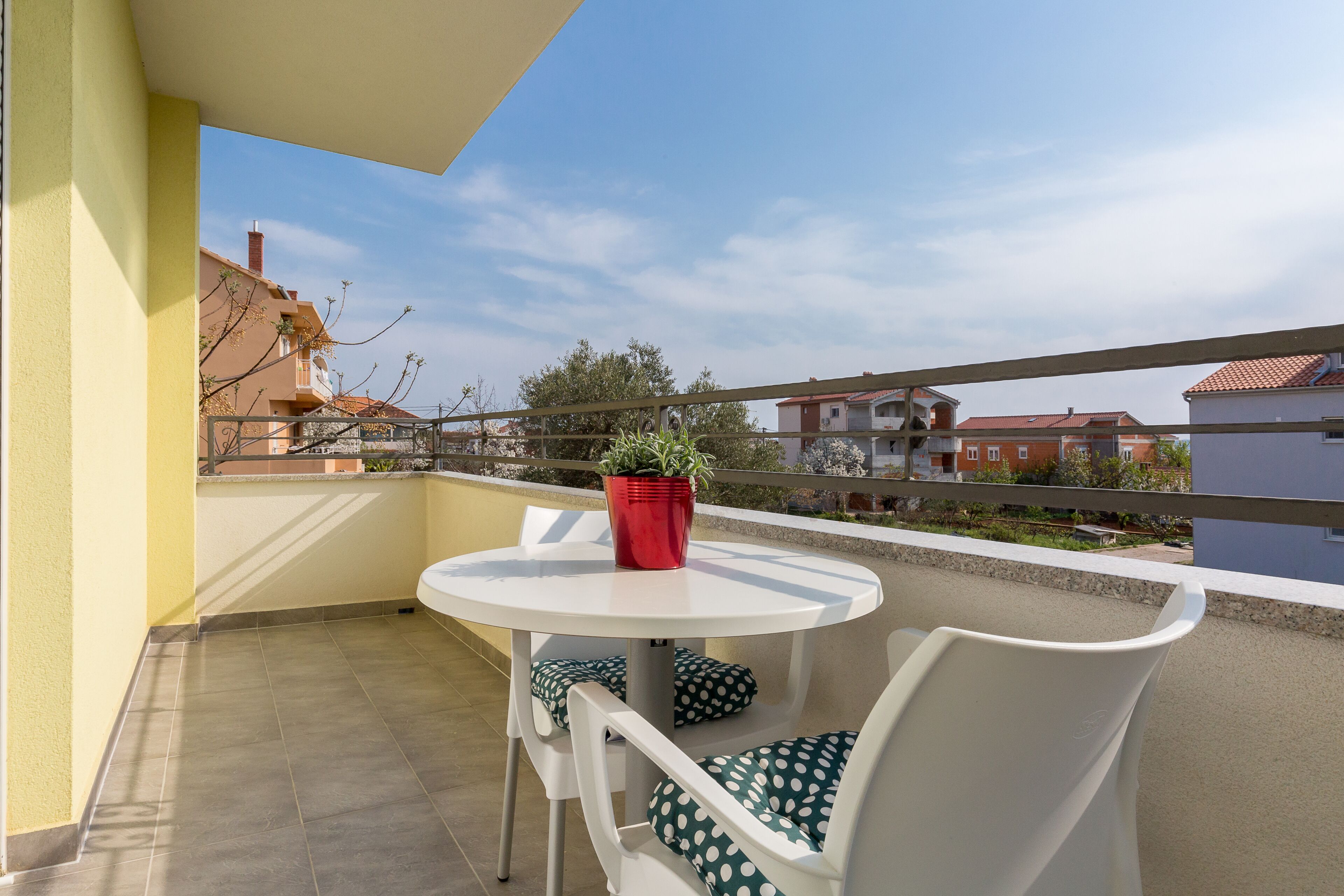 A balcony with a round table and two chairs, a red pot on the table, and a view of buildings in the background.