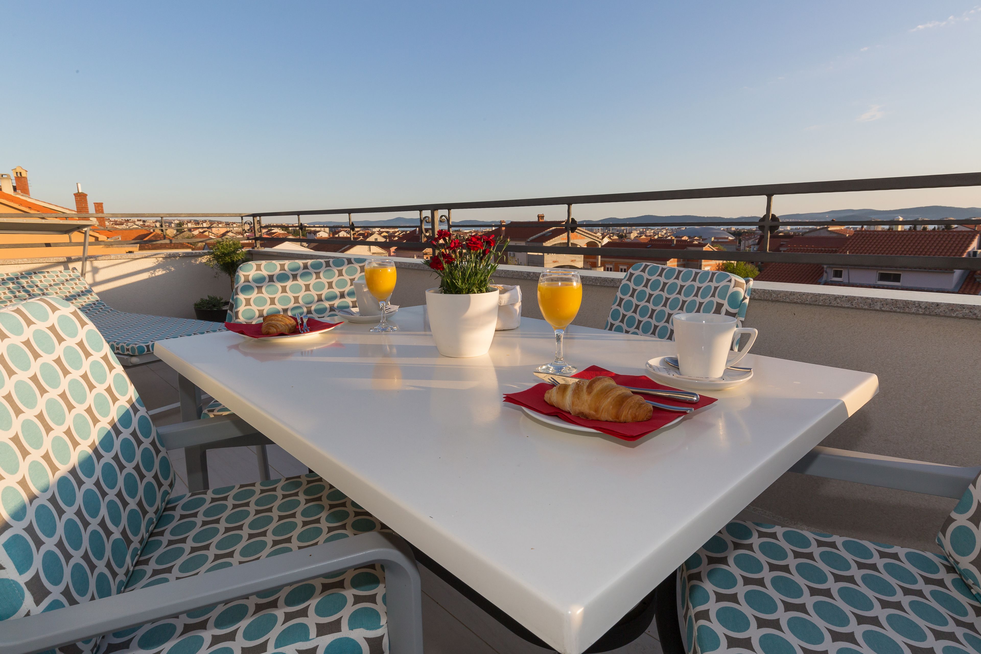 Terraza en la azotea con una mesa preparada para el desayuno, con cruasanes, zumo de naranja y un jarrón de flores.
