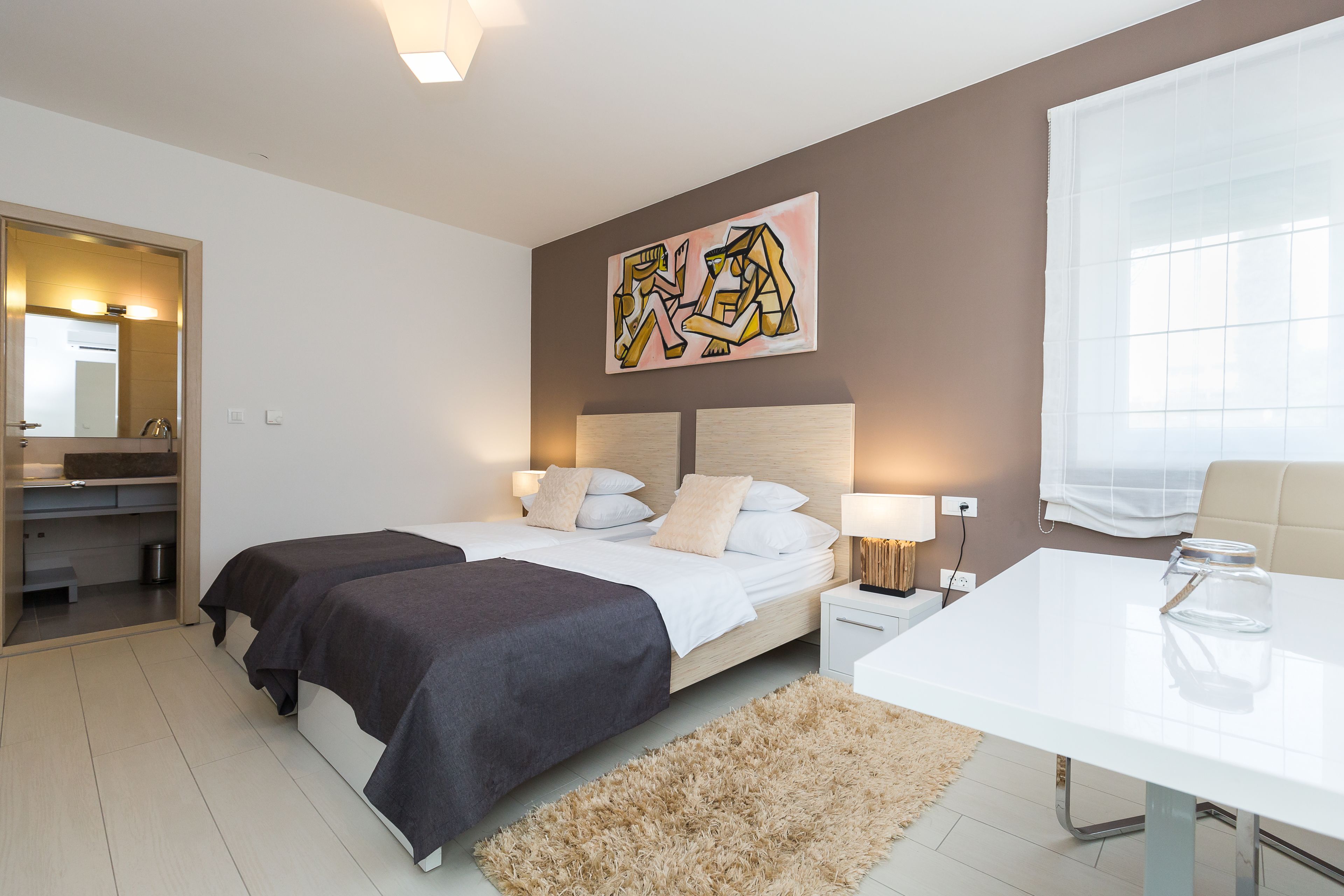 Ein modernes Schlafzimmer mit zwei Betten, einem Nachttisch mit Lampe, einem großen Fenster mit durchscheinenden Vorhängen und einem Wandbild.