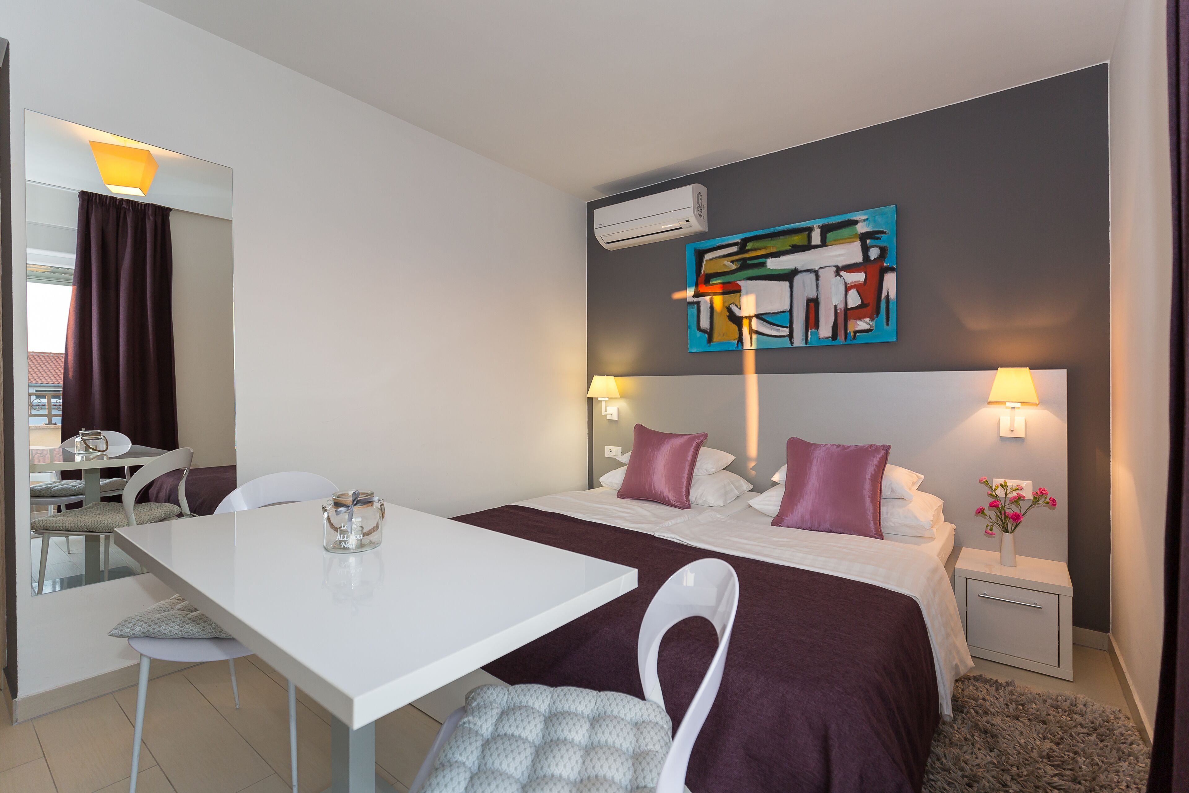 A hotel room with a bed, bedside tables, a desk, chairs, and a painting on the wall.