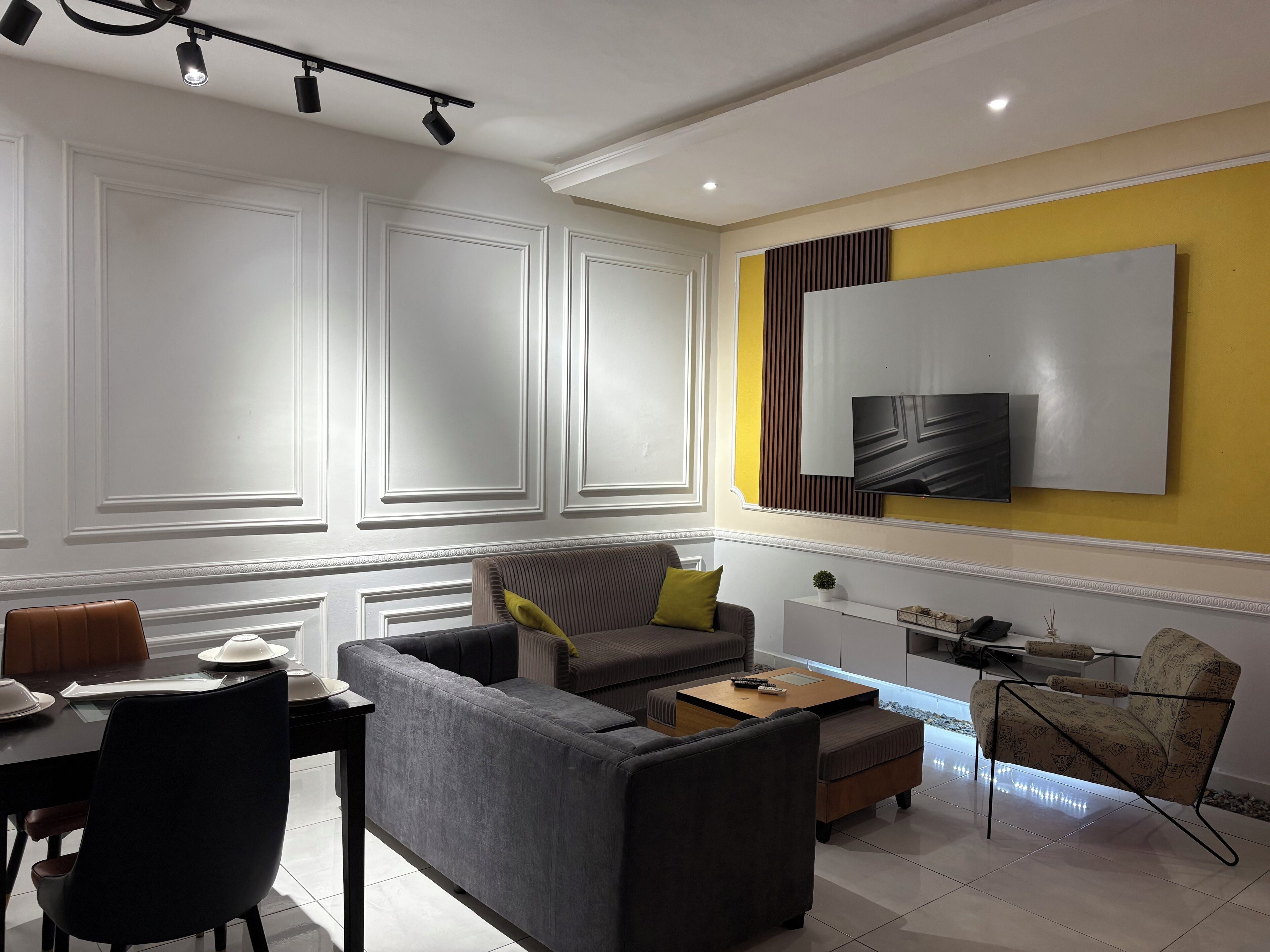 Una sala moderna con un sofá gris, una mesita de centro, una mesa de comedor con sillas y un televisor montado en una pared amarilla.