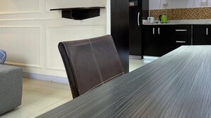 Un comedor moderno con una mesa larga, un sillón de cuero marrón, un espejo redondo y una zona de cocina con una campana extractora negra.