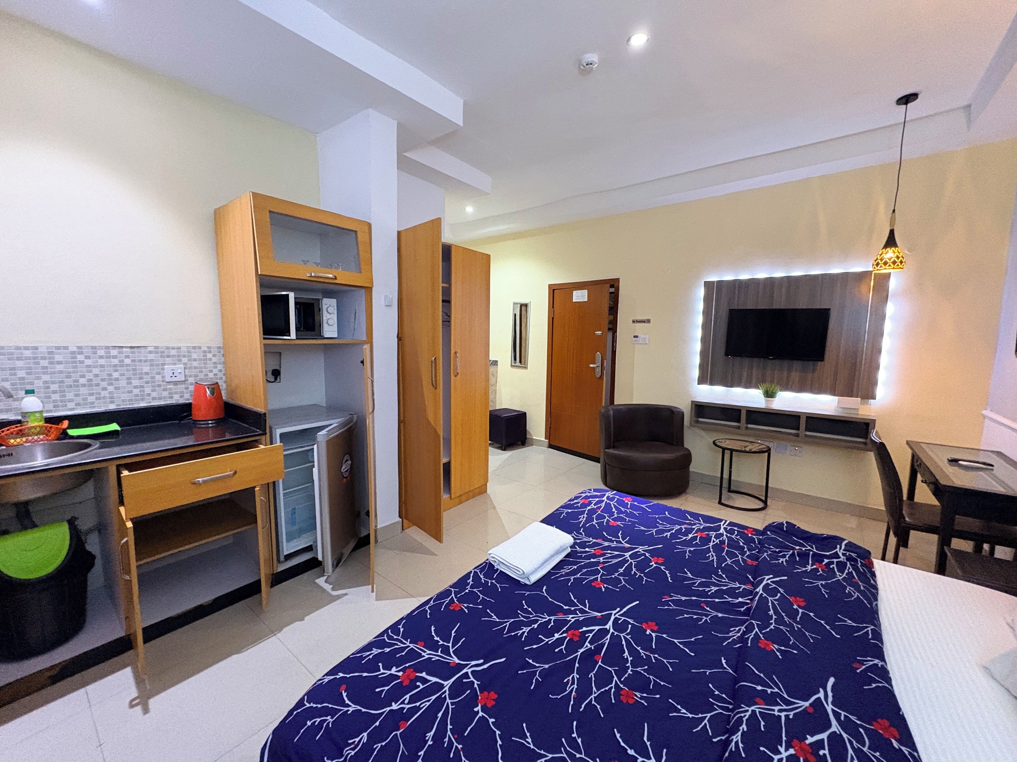 A compact hotel room with a kitchenette, a bed with a floral bedspread, a seating area with a chair and table, and a TV mounted on the wall.
