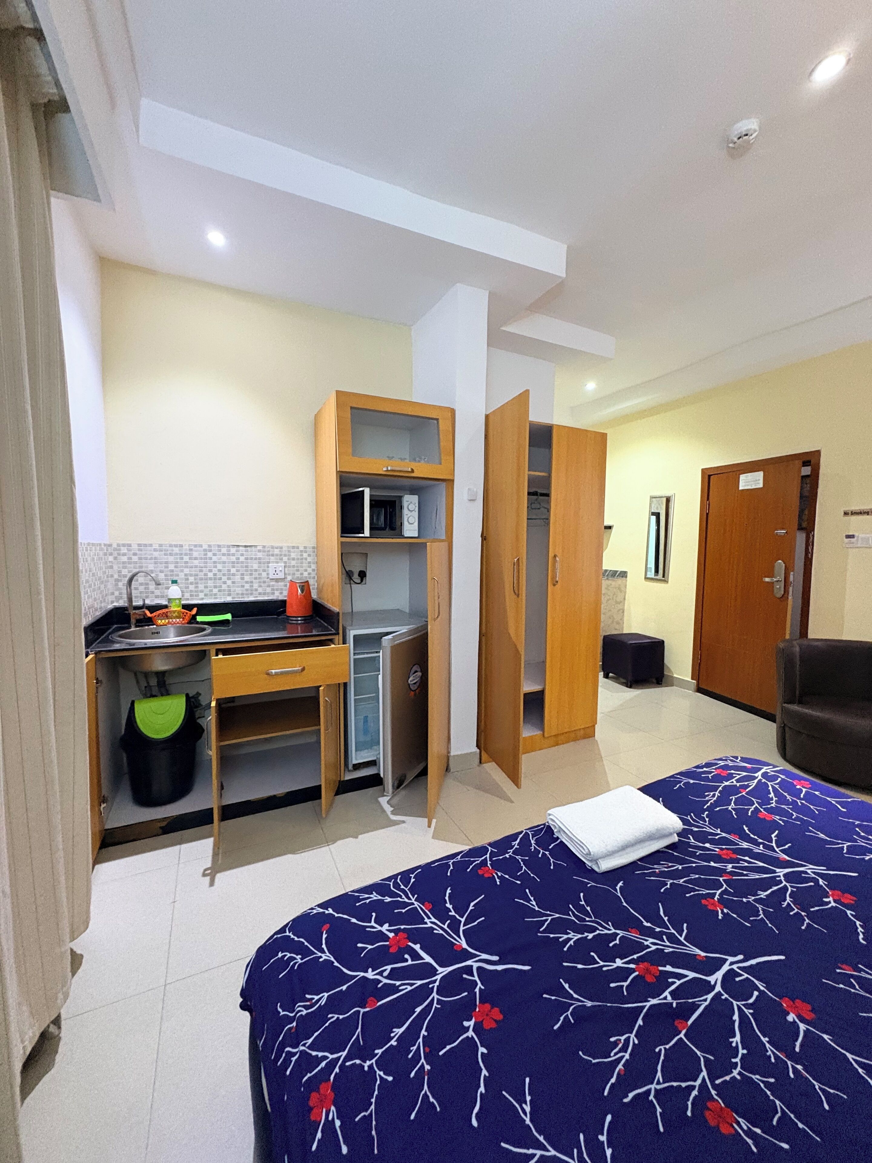 A compact hotel room with a bed, a kitchenette, a wardrobe, and a seating area.