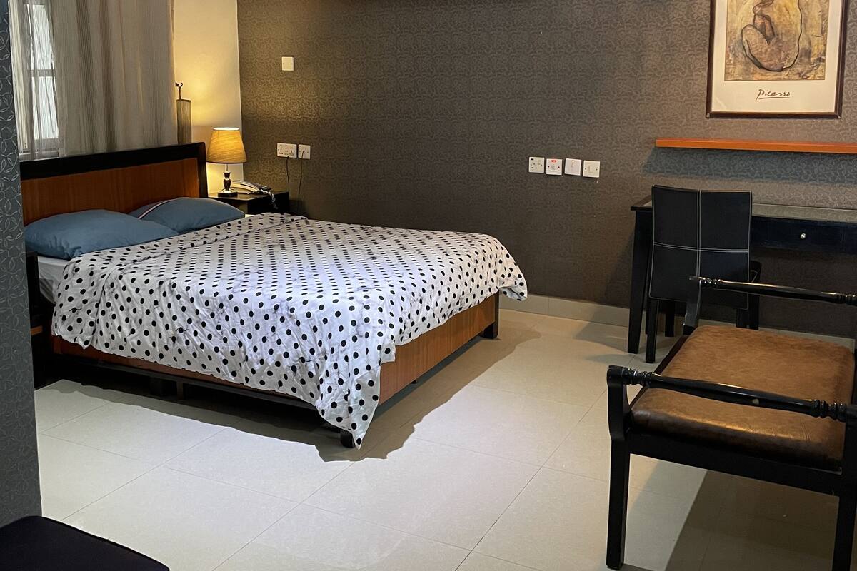 A modern hotel room with a bed, a desk, a chair, a wall-mounted air conditioner, a framed artwork, and a window with curtains.