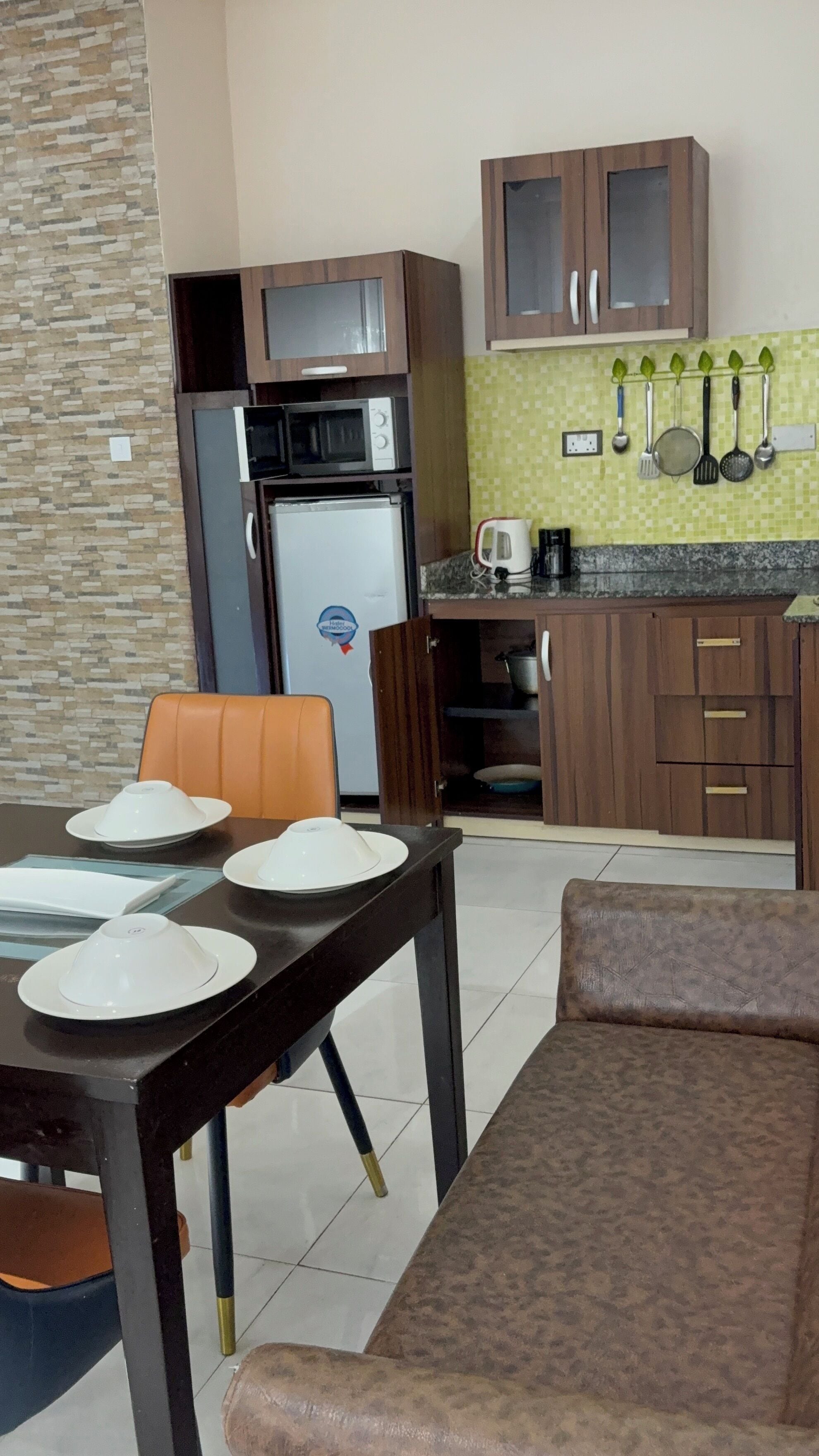 A compact kitchen with a microwave, refrigerator, and a coffee maker. There is a dining table with plates and a chair. The kitchen has wooden cabinets and a tiled backsplash.