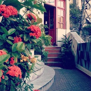 A building with a red door and steps leading to a white door. There are red flowers and green plants around the entrance. - Mambembe Hostel (Rio de Janeiro)