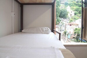 A single bed with a white mattress and pillow in a room with a view of a residential area through an open window.